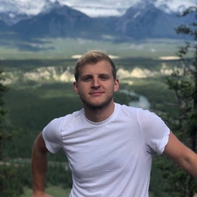 Portrait of Sam Frenzel. He's wearing a white t-shirt with the view of the mountains behind him.